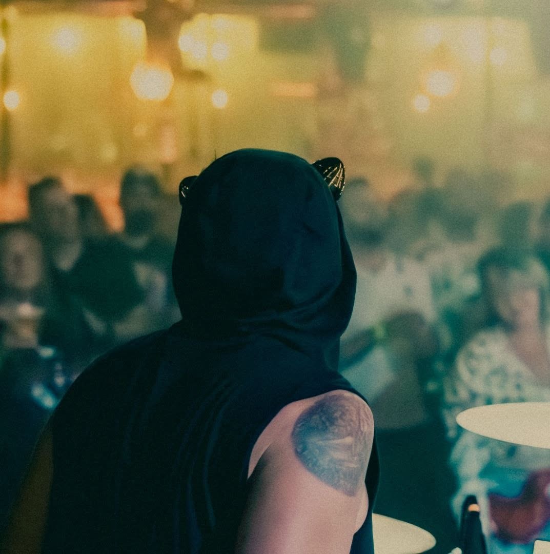 A hooded band member at the drums, with a warmly lit crowd beyond